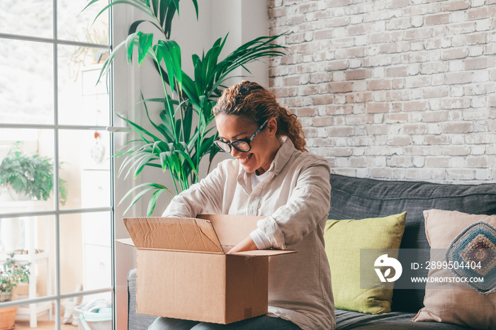 Woman unpacking delivdery box opening package at home. Happy young lady looking at carton box while sitting on sofa in living room at modern apartment. Caucasian female checking out delivered stuff.