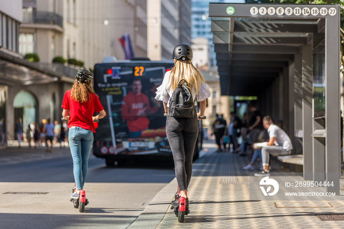 Rear view of trendy fashinable teenager girls riding public rental electric scooters in urban city environment. New eco-friendly modern public city transport in Ljubljana, Slovenia.