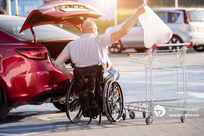 Adult disabled man in a wheelchair puts purchases in the trunk of a car in a supermarket parking lot