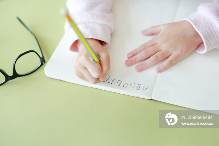 Close-up hands of little child writes ABC in a book or notebook with pencil on table in classroom against white background.