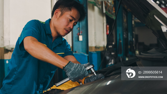 Professional car mechanic screwing details of car engine on lifted automobile at repair service station. Skillful Asian guy in uniform fixing car at mechanics garage at night.