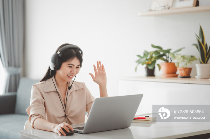 Smiling Asian woman using headset having video call meeting with colleague via laptop at home in living room. Work from home and self quarantine during coronavirus pandemic