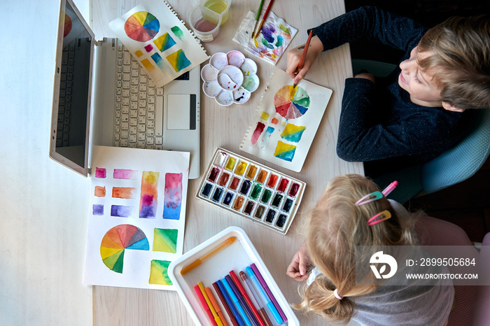 Children painting pictures with watercolor paints during art lesson. Pupils are concentrating on drawing with brush. Watercolor color wheel and palette. Color theory beginner hobby lessons