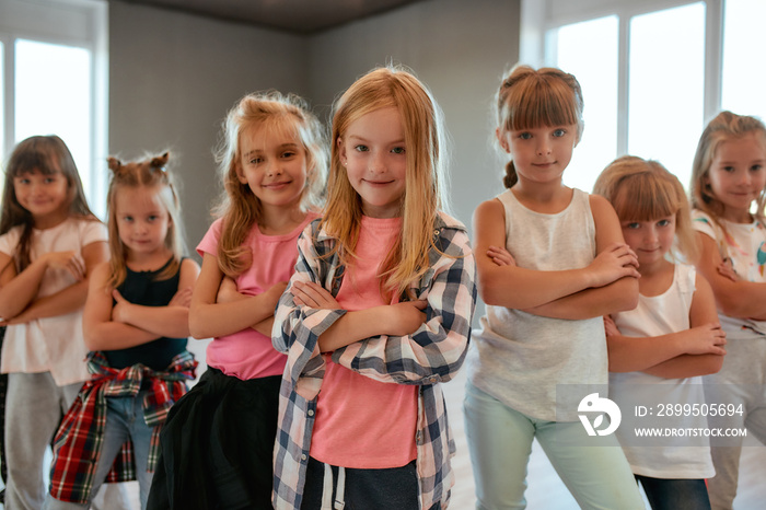 Cool dancers. Portrait of positive little girls keeping arms crossed and smiling at camera while standing in the dance studio. Choreography class