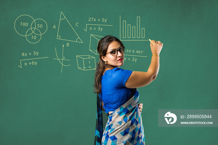 Portrait of asian Indian lady teacher in saree stands against writing board conducting online class