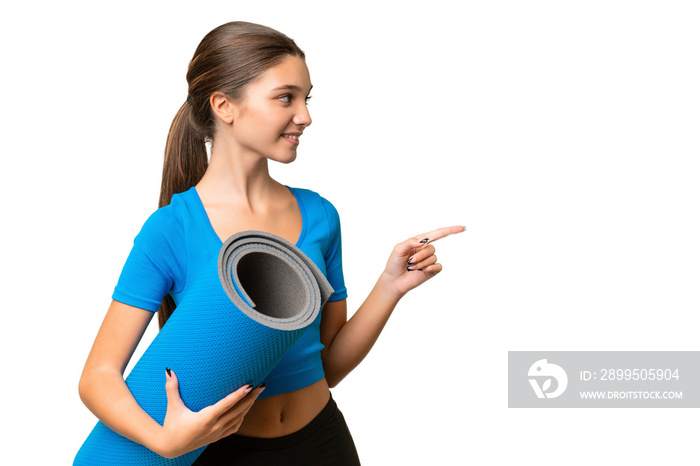 Teenager caucasian girl going to yoga classes while holding a mat over isolated background pointing to the side to present a product