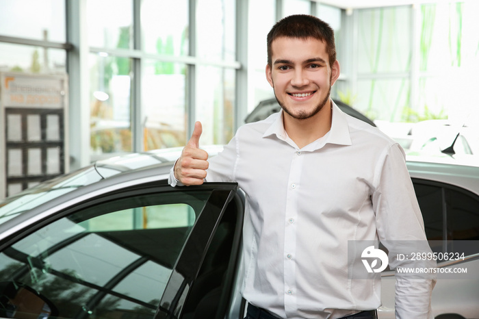 Handsome car salesman showing thumb up gesture near automobile in dealership centre