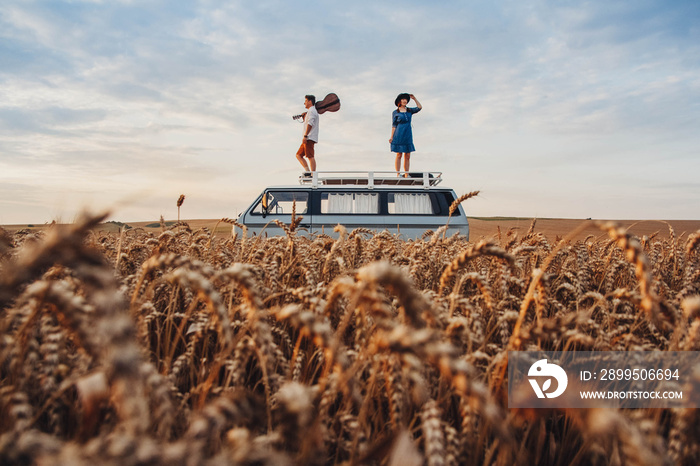 Man with a guitar and woman in a hat are standing on the roof of a car in a wheat field