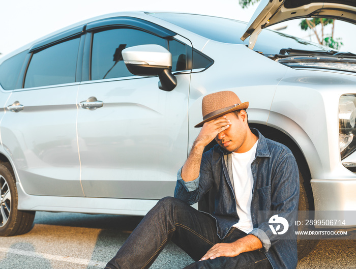 A young Asian tourist has driven a car and broke down on the way. He sat down on the floor beside the wheel of a car.