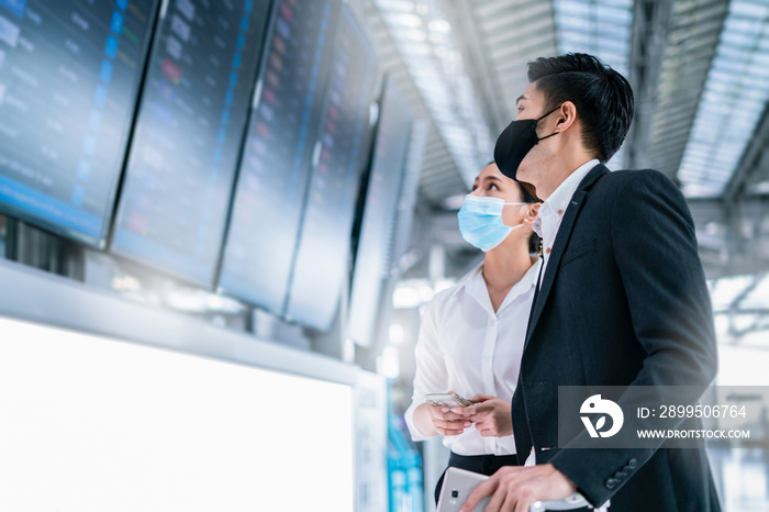 Smart asian businessman and businesswoman wear mask protection in airport terminal,discuss and planning business strategy while waiting flight on information board background.Partner meeting concept.