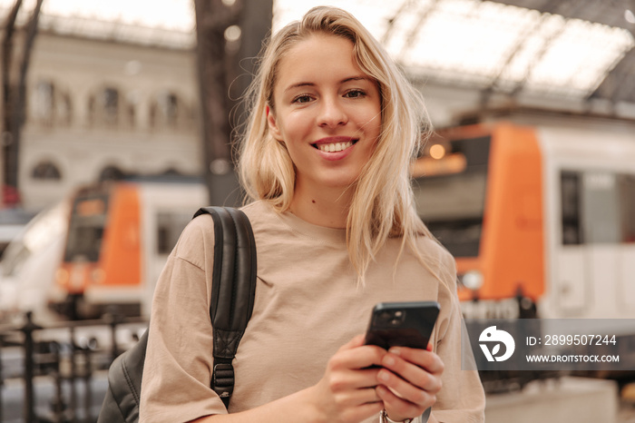 Portrait of beautiful young woman on the background of railway station . Focused on smiling blonde in brown t-shirt with phone