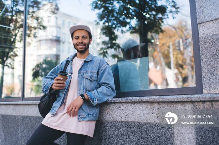 Half length of prosperous male tourist in stylish denim jacket smiling at camera during leisure daytime in city, happy Hispanic hipster guy with takeaway coffee to go enjoying travel getaway trip