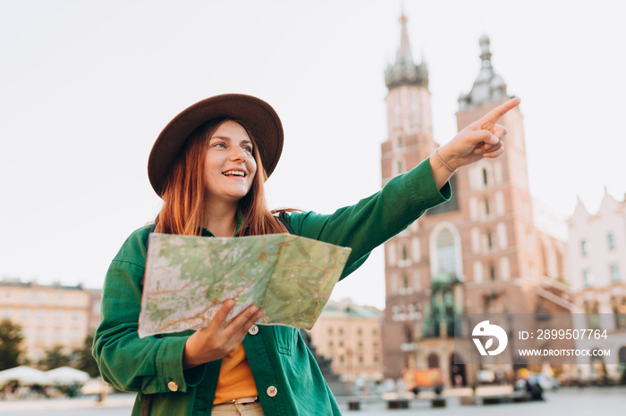 Attractive female tourist is exploring new city. Redhead woman pointing finger and holding a paper map on Market Square in Krakow. Traveling Europe in autumn. St. Marys Basilica. Active lifestyle