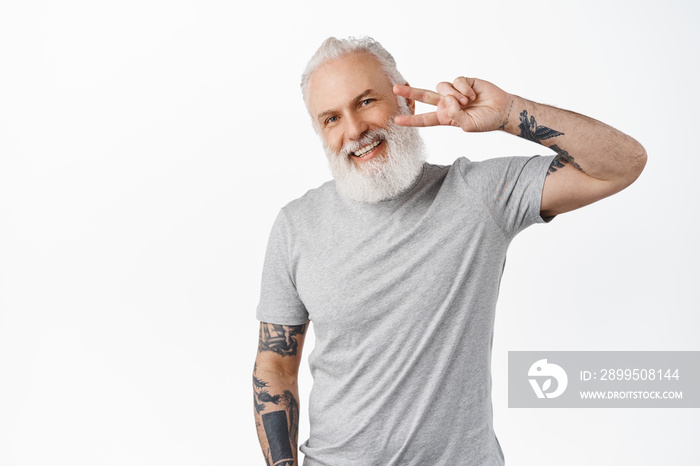 Happy cute senior guy with tattoos and beard, shows peace v-sign, looking kawaii and smiling at camera, standing in grey casual t-shirt against white background