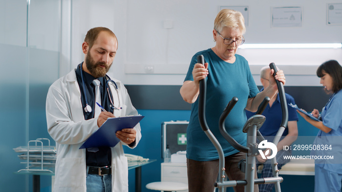 Physician giving advice to elder woman on stationary bike, using alternative medicine to treat physical injury. Specialist assisting patient to do recovery procedure and increase mobility.