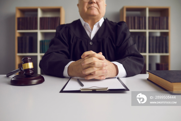 Serious fair incorruptible old male judge at table with gavel and case papers in courtroom. Honest wise senior lawyer in uniform sitting with clasped hands at desk. Concept of court of law and justice