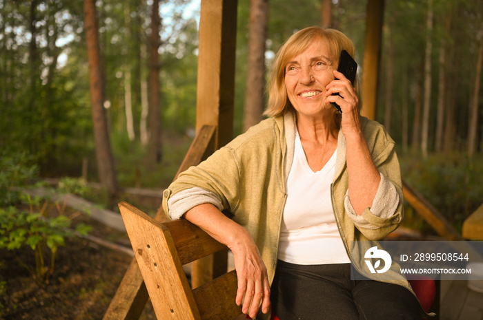 Technology, old age people concept - elderly senior old happy smiling woman speaks cellphone smartphone outdoor at wooden terrace in the back yard summer garden countryside.