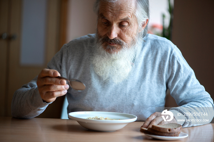 Old sad man with a long gray beard sitting by the table and eating soup and bread