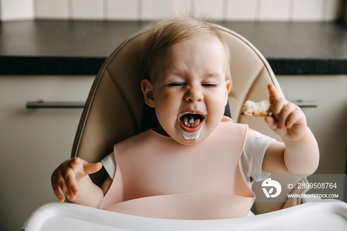 10 months old baby sitting in high chair, eating bread and sour cream, grimacing.