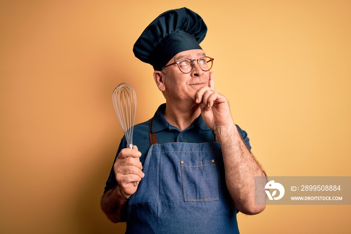 Middle age hoary cooker man wearing apron and hat holding whisk over yellow background serious face thinking about question, very confused idea