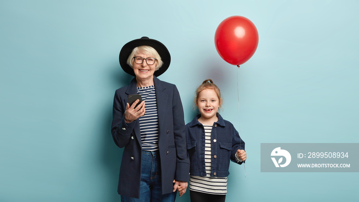 Image of cheerful grey haired woman in hat, holds mobile phone, sends text message, keeps hand of little granddaughter who carries red balloon, isolated over blue background. Family and celebration