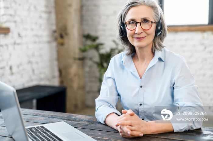 Mature senior office worker businesswoman with a pleasant smile in headset and glasses looking at the camera, sitting at the desk. Female call center operator is feeling motivated and ready to work