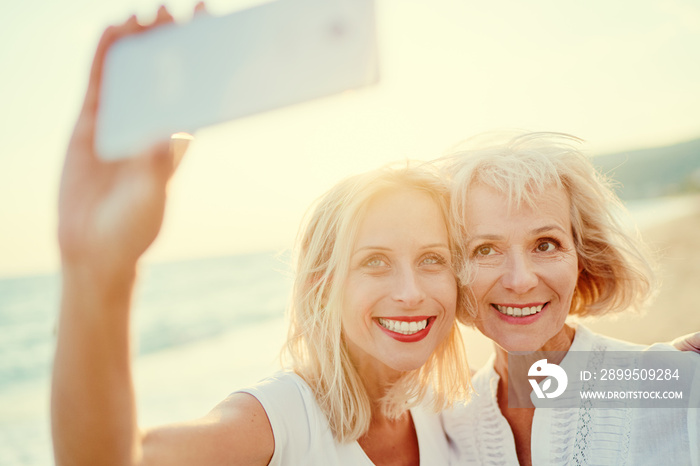 Outdoor portrait of smiling happy caucasian senior mother with her adult daughter taking selfie on smartphone on sea beach.