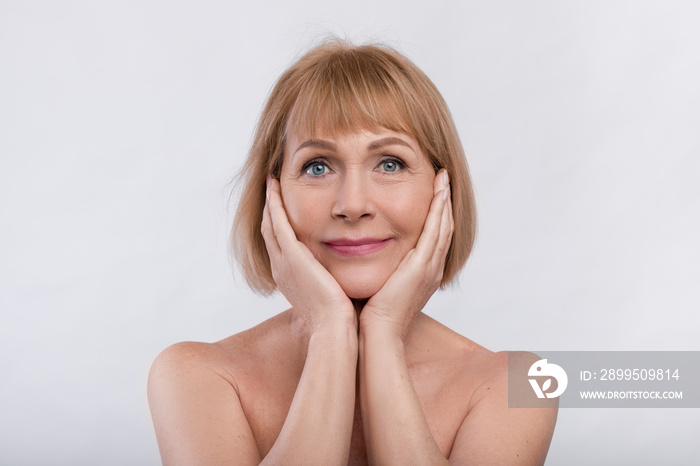 Portrait of charming nude senior lady touching her face over light studio background