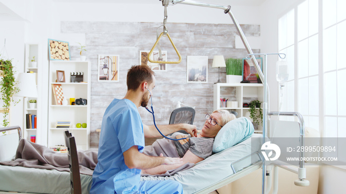 Male nurse checking the heartbeats of an sick old lady lying in hospital bed in bright and cozy nursing home