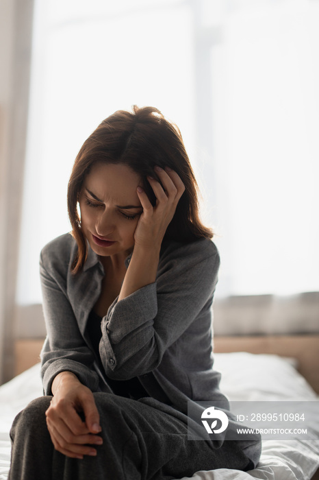 Brunette woman sitting on bed, suffering from pain and touching head at home