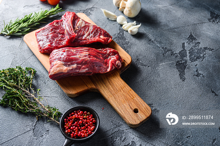 Raw flap steak flank cut with Machete, Skirt Steak, on woods chopping board, with herbs tomatoes peppercorns over grey stone surface background  side view space for text