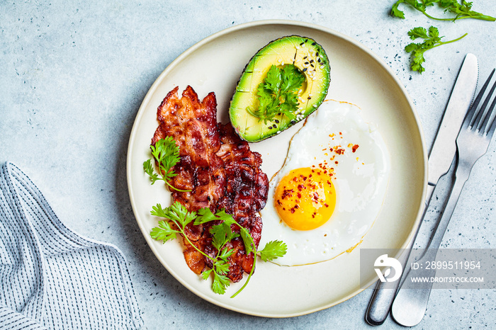 Keto breakfast - fried egg, avocado and fried bacon in white plate, top view. Keto diet concept.