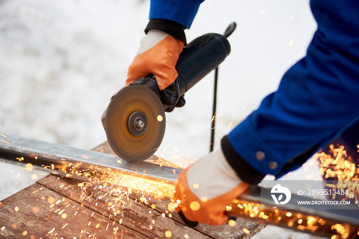 Cropped close up of an industrial worker in protective uniform and gloves cutting metal sawing welding iron outdoors in winter factory industry metalworking electric sparks steel profession.