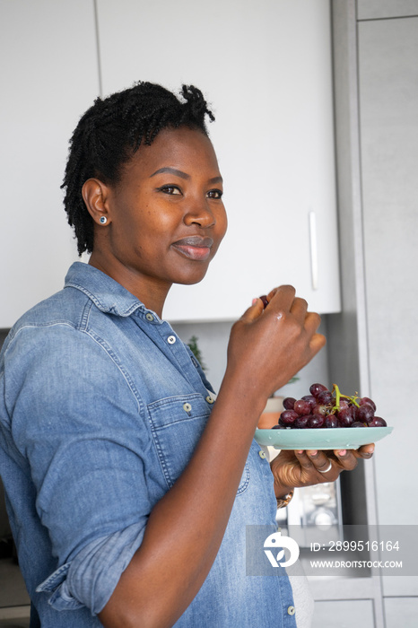 Smiling pregnant woman holding plate with red grapes in kitchen
