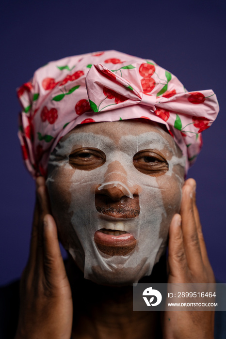 Portrait of man in hair cap applying facial mask against purple background