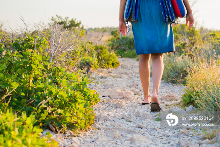 Close up shot of young woman legs wearing flip flops and summer clothes carrying beach sleeping pad while walking on a trail. Summer holidays and travel concept.