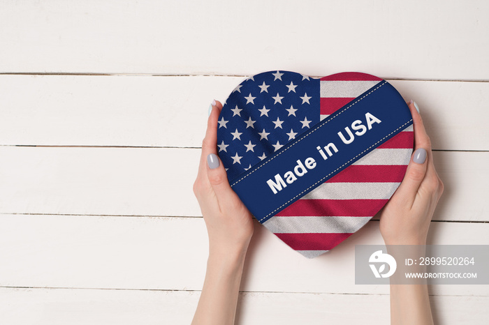 Inscription Made in USA, the flag of USA. Female hands holding a heart shaped box. White wooden table on a background. Place for text