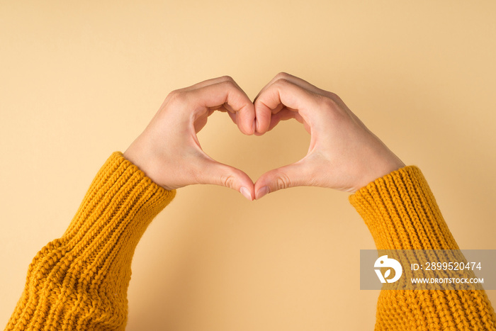 First person photo of woman’s hands in yellow sweater making heart with fingers on isolated pastel orange background