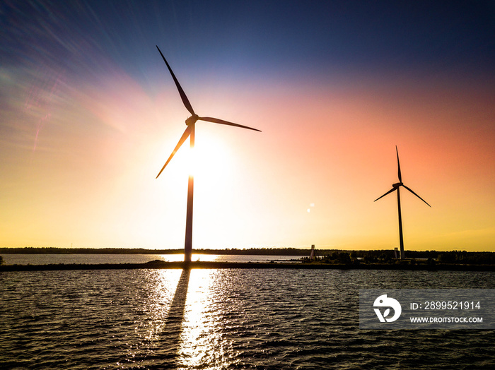 aerial photo from drone of two windmills standing at sea pier with bright colored sunset behind them, shadows are seen on wavy sea surface