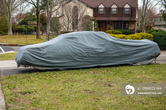 Historic antique muscle car in the driveway of a suburban house completely covered with a gray car protection cover