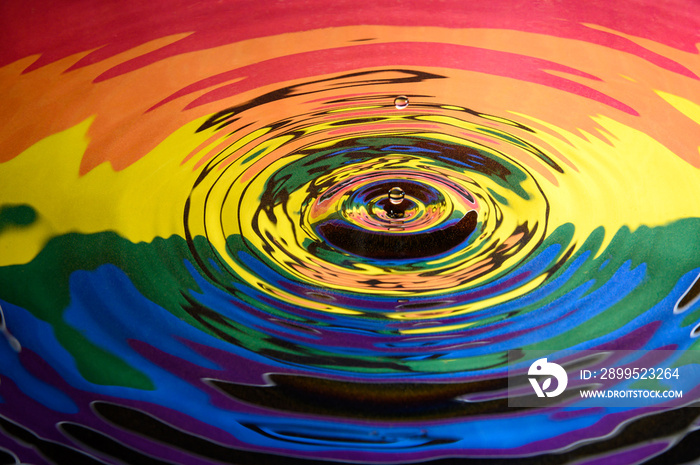 The Gay Pride Extinction Rebellion flag and logo reflected in circular waves from a water splash along with two falling droplets of water