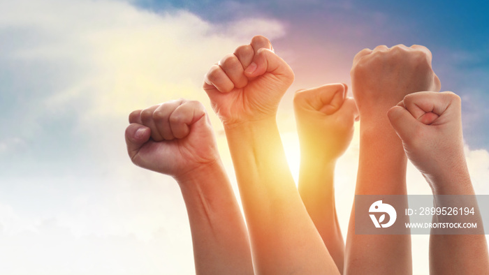 Rising fist of adult people, male and female, dramatic blue sky with sun light, anger protest revolution inspiration motivation or teamwork concept