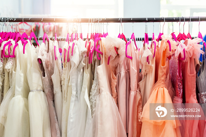 Rack with many beautiful holiday dresses apparel on hangers at female fashion showroom indoor. Kid girl dress hire studio for celebration birthday party or photography session event. Bright sun day