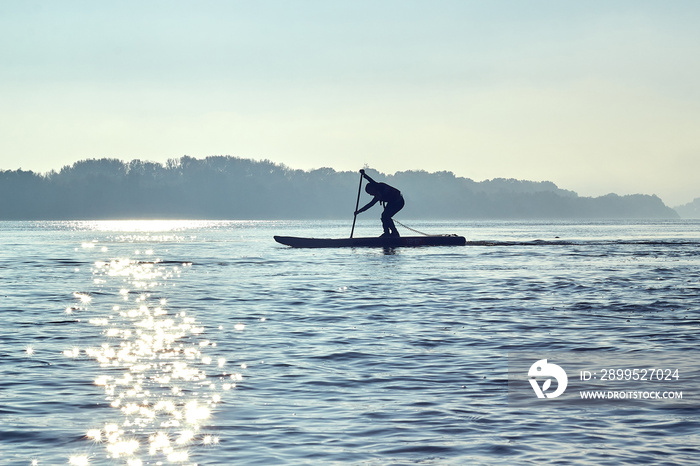 Silhouette of teenager rowing on SUP (stand up paddle board) at sunrise in a haze at Danube river