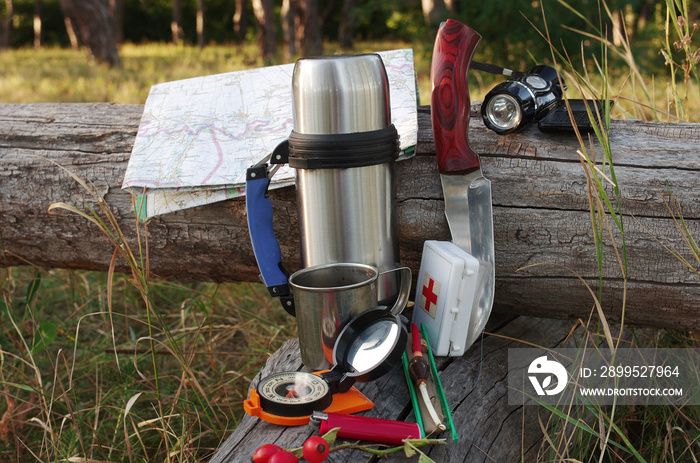 A whale of items for survival on a natural background. A knife, a thermos, a mug, a compass, a map, a lantern, matches, a first aid kit, a fishing rod.