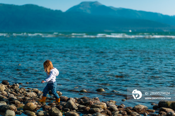 A young girl in rain boots walks along the rocky shore in front of a mountain in Alaska