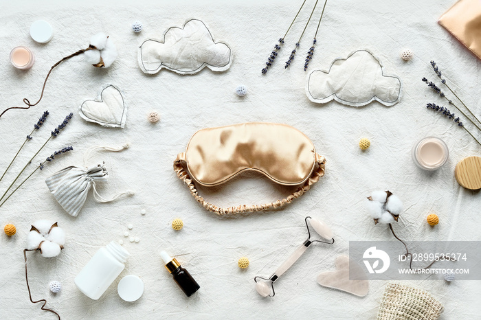 Healthy night sleep flat lay. Pale yellow sleep mask, face stone roller, Gua Sha stone and essential oil. Sleeping pills, night cream. Off white textile bed sheet. Dry lavender, cotton flowers.