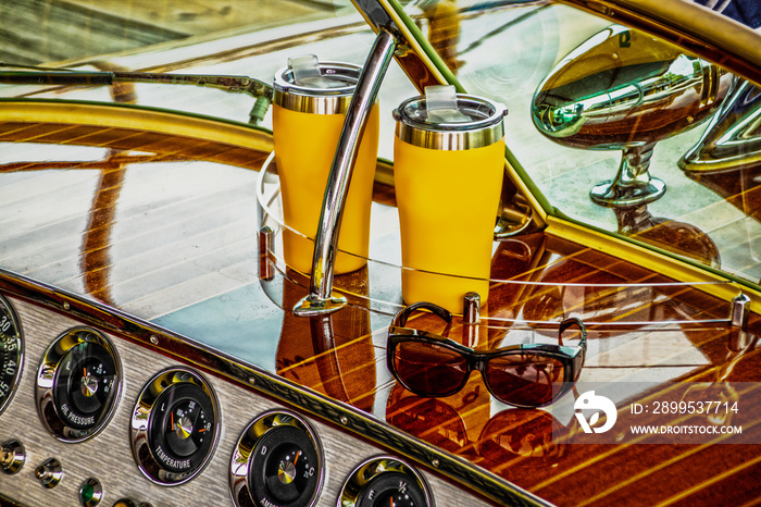 Dash of antique classic wooden speedboat holding two insulated drink cups and sunglasses and gauges below - very shinny with reflections on wood and windshield - closeup