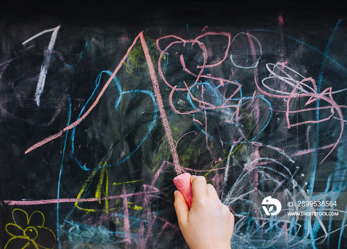 Little girl child draws with her right hand with pink chalk on a black wooden board, easel a picture, scribbles and a straight line. Drawing lesson at school.