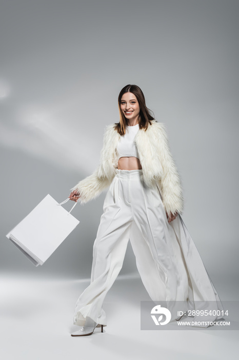 Cheerful young woman in faux fur jacket smiling at camera and holding shopping bags on abstract grey background.